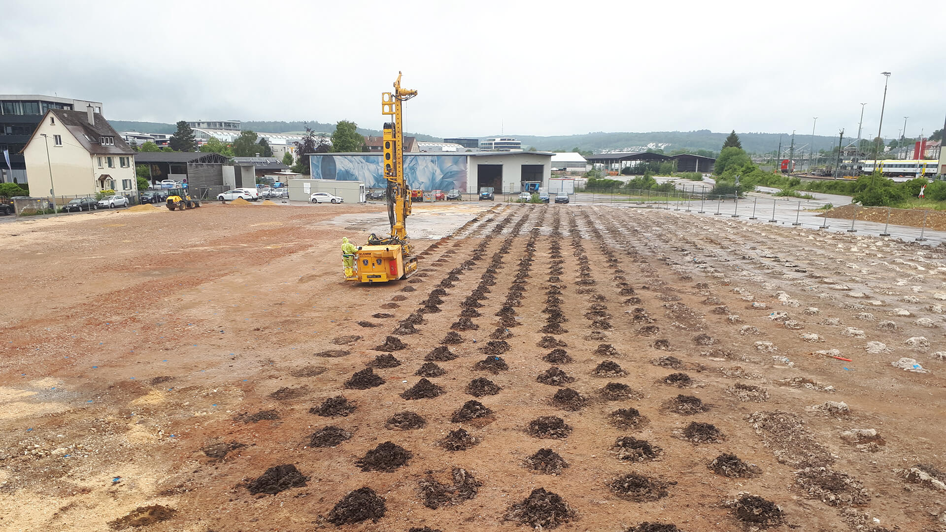 Großflächige Baustelle mit Raster aus Bohrlöchern und Erdaushub zur Bodensanierung; im Vordergrund ein Bohrgerät, im Hintergrund Gewerbebauten und Infrastruktur bei bewölktem Himmel.