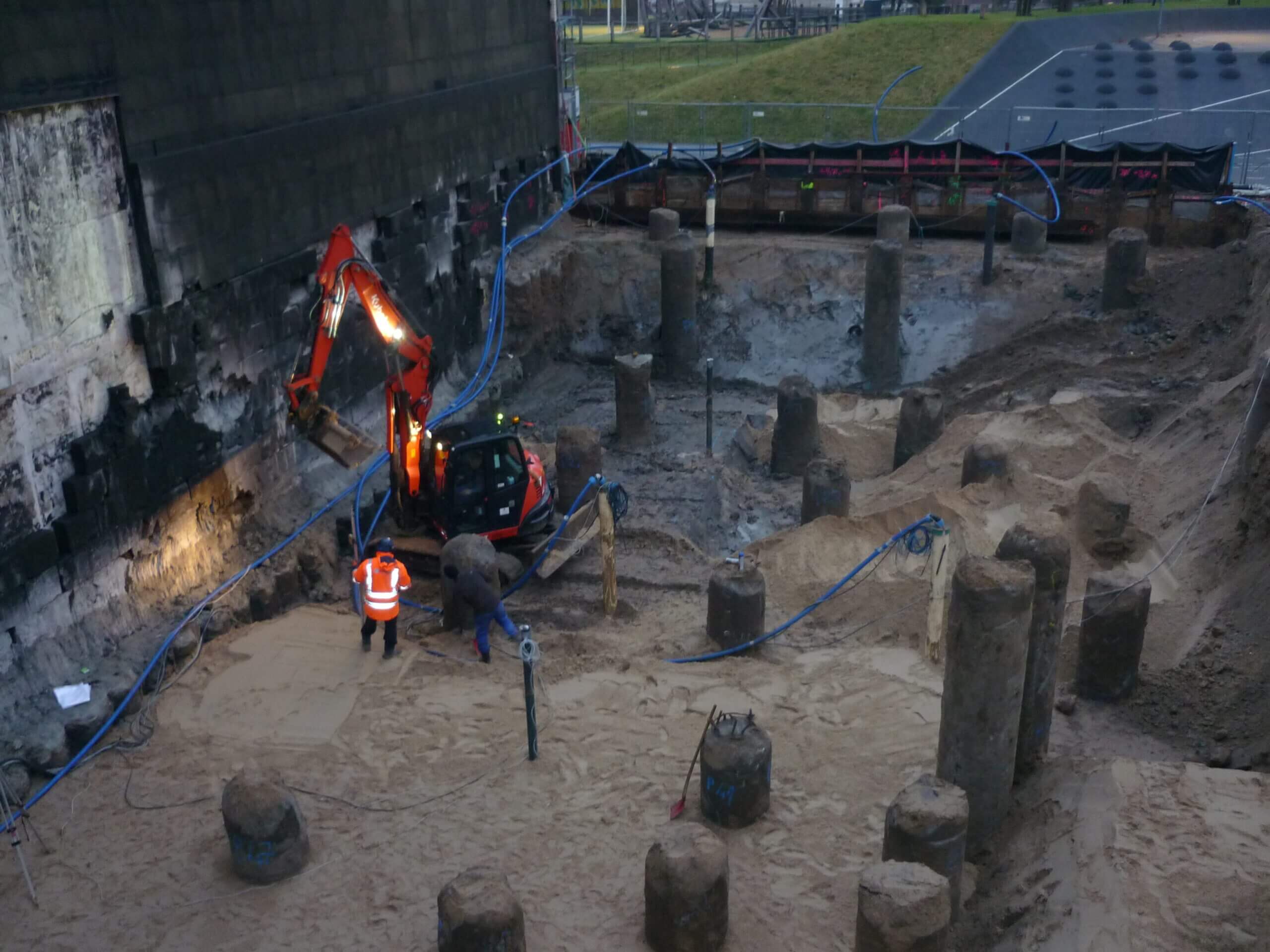 Tiefe Baugrube mit freigelegten Pfählen und sandigem Untergrund; ein Bagger arbeitet zwischen den Pfählen, zwei Bauarbeiter überwachen die Arbeiten, Schläuche zur Wasserhaltung verlaufen durch die Baugrube.