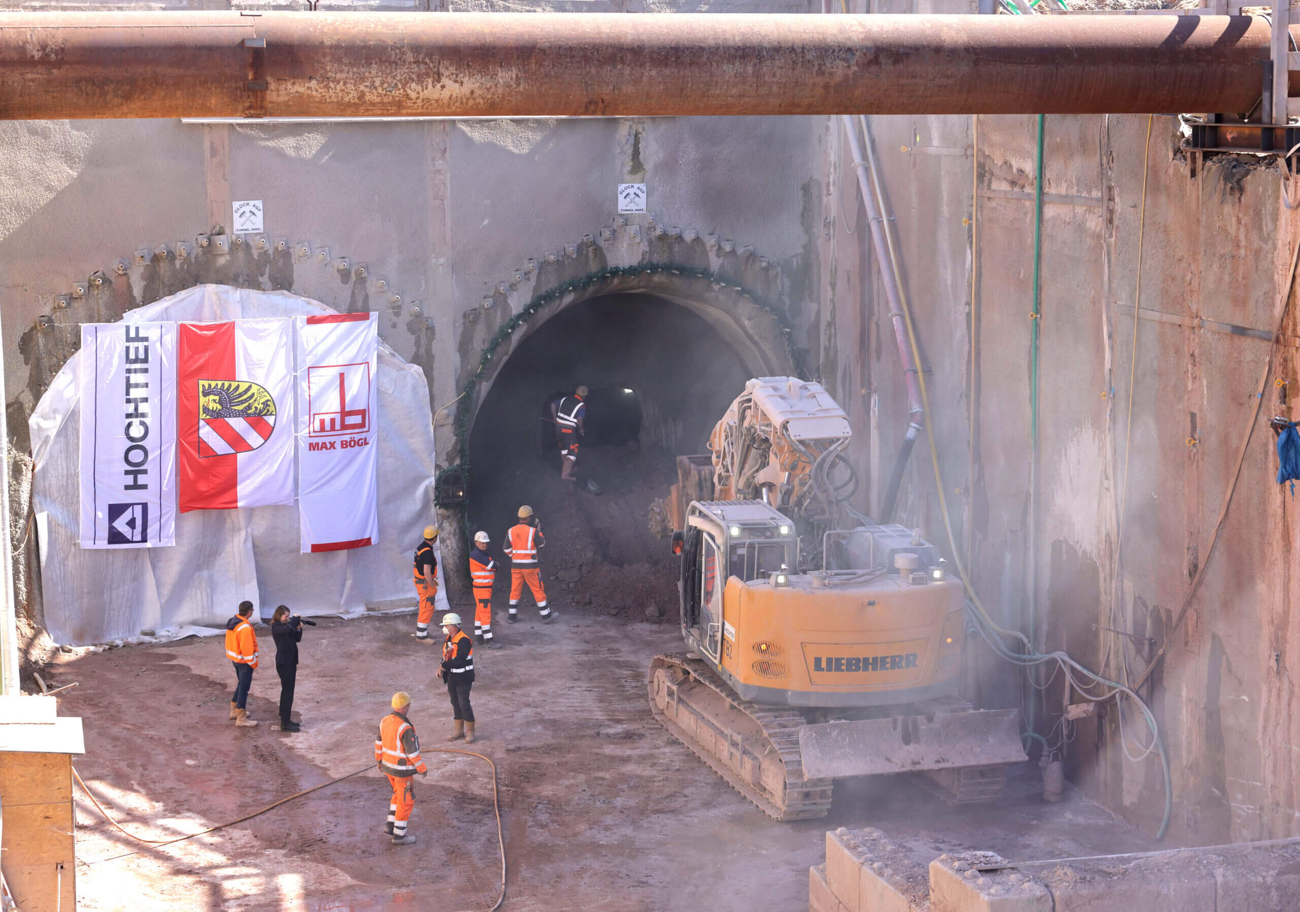 Tunnelbaustelle mit feierlichem Durchbruch: Bauarbeiter in Warnkleidung stehen vor der Tunnelöffnung, ein Bagger räumt Material ab; Banner der beteiligten Bauunternehmen hängen am Tunneleingang.